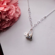 A silver pendant chain featuring a geometric heart-shaped bag of treasure design, displayed on a white surface with pink flower accents in the background.